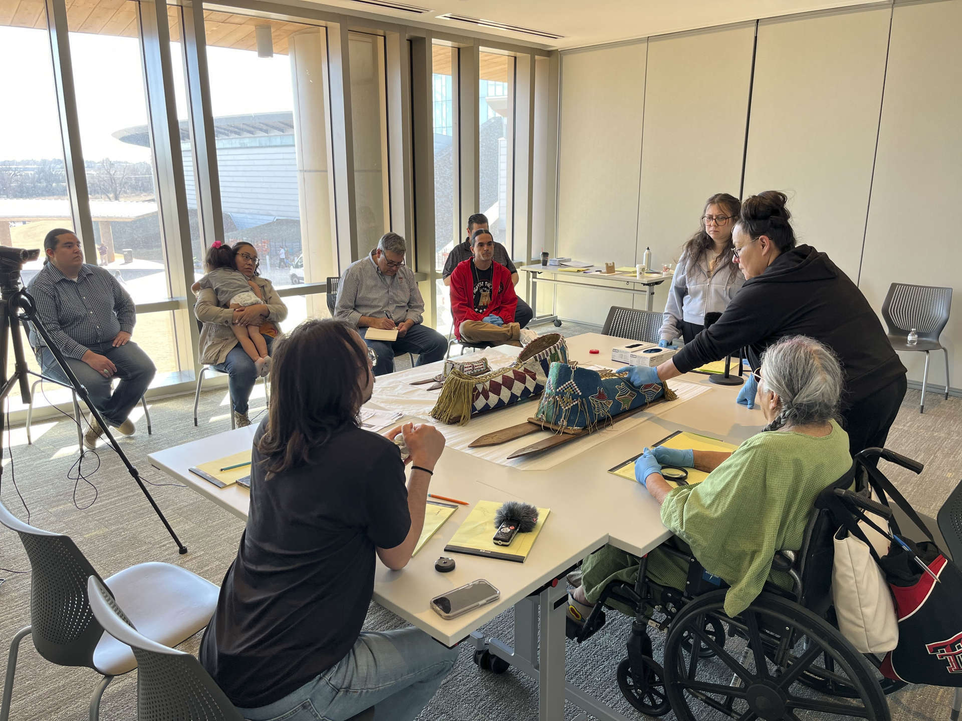 Seven people around a large table examining two Kiowa beaded cradleboards. 