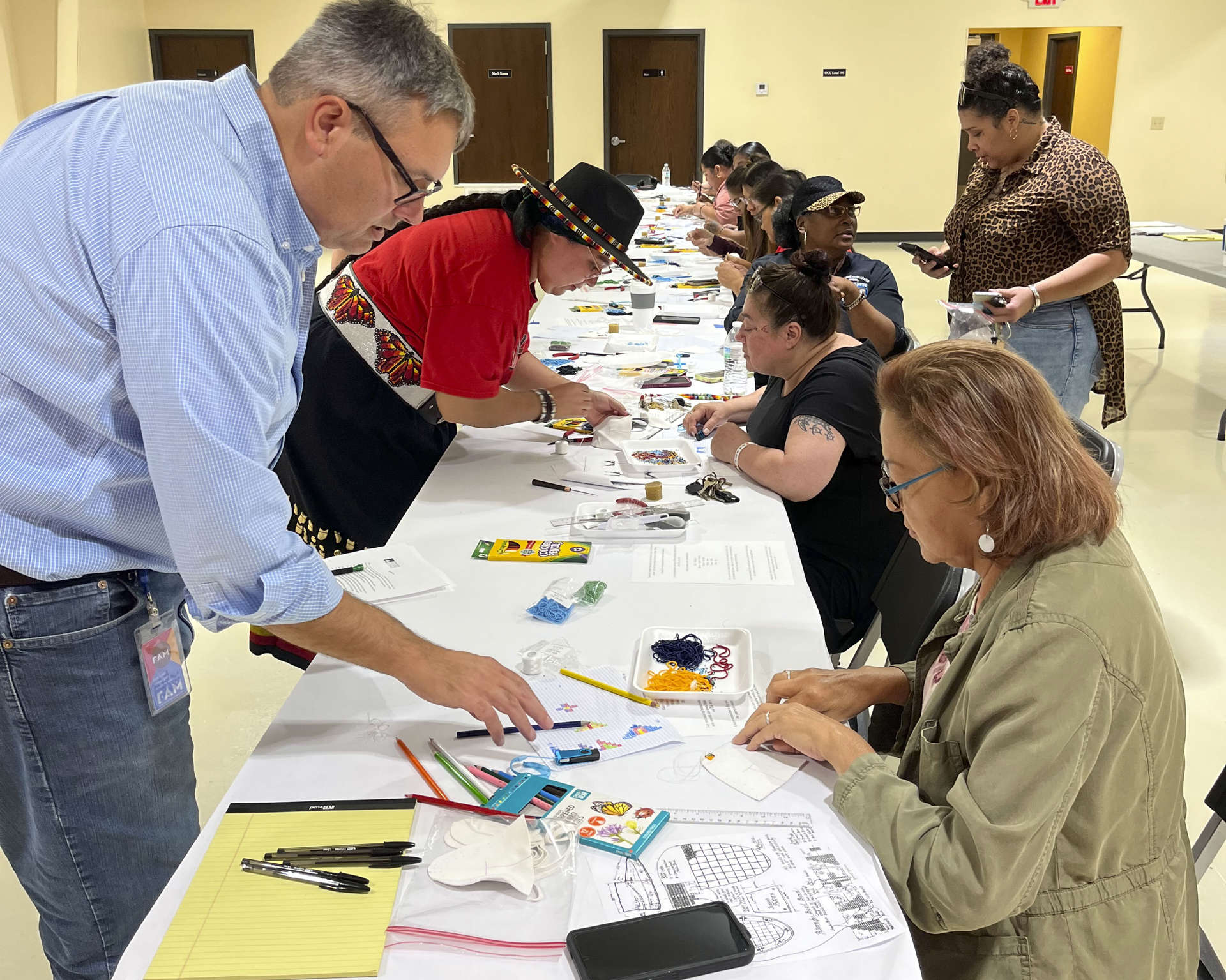 Beading class participants sitting at a long table and two people standing on the opposite side. 