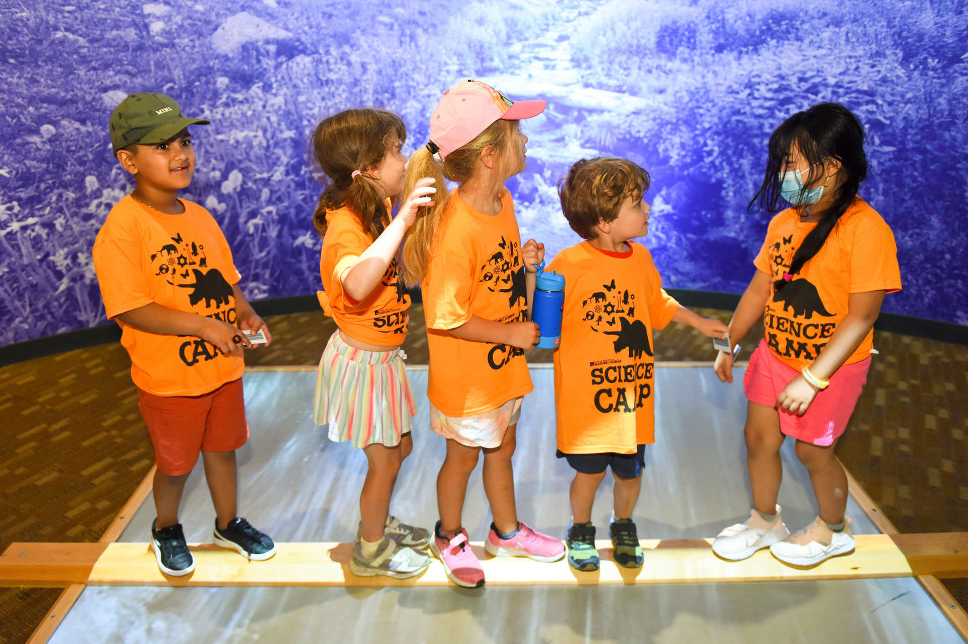 A group of children in orange shirts stands together on a wooden platform, smiling and enjoying their time in Expedition Health. .