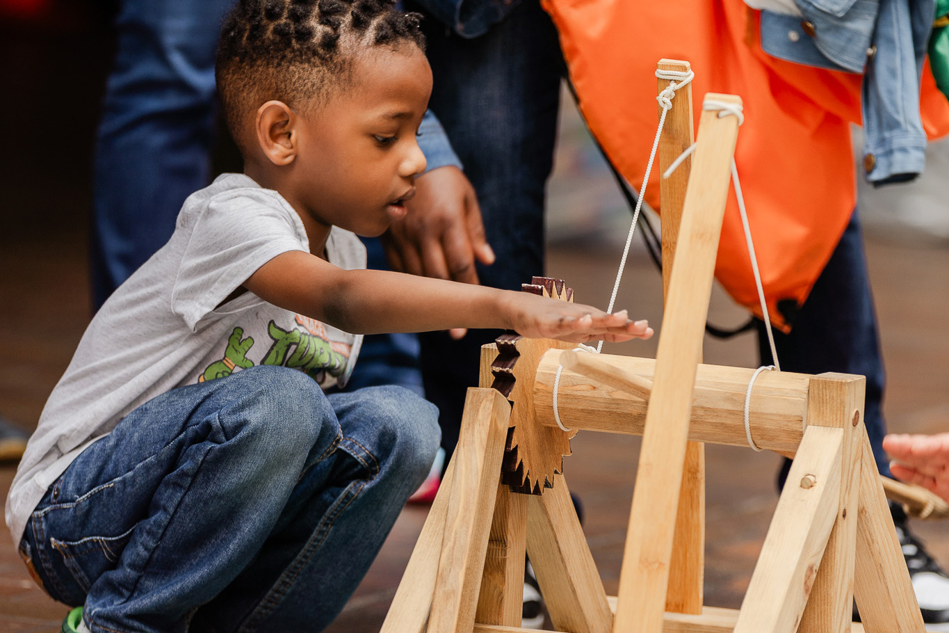 A child operates a wooden mechanical apparatus during an interactive hands-on program, surrounded by adults and other visitors.