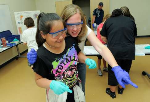A student and adult wearing gloves smile while pointing at an object out of frame