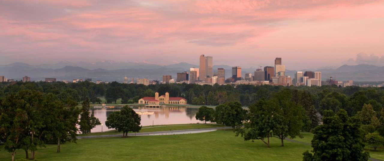 A view of City Park and Downtown Denver including a lake, the skyline, and mountains at sunset